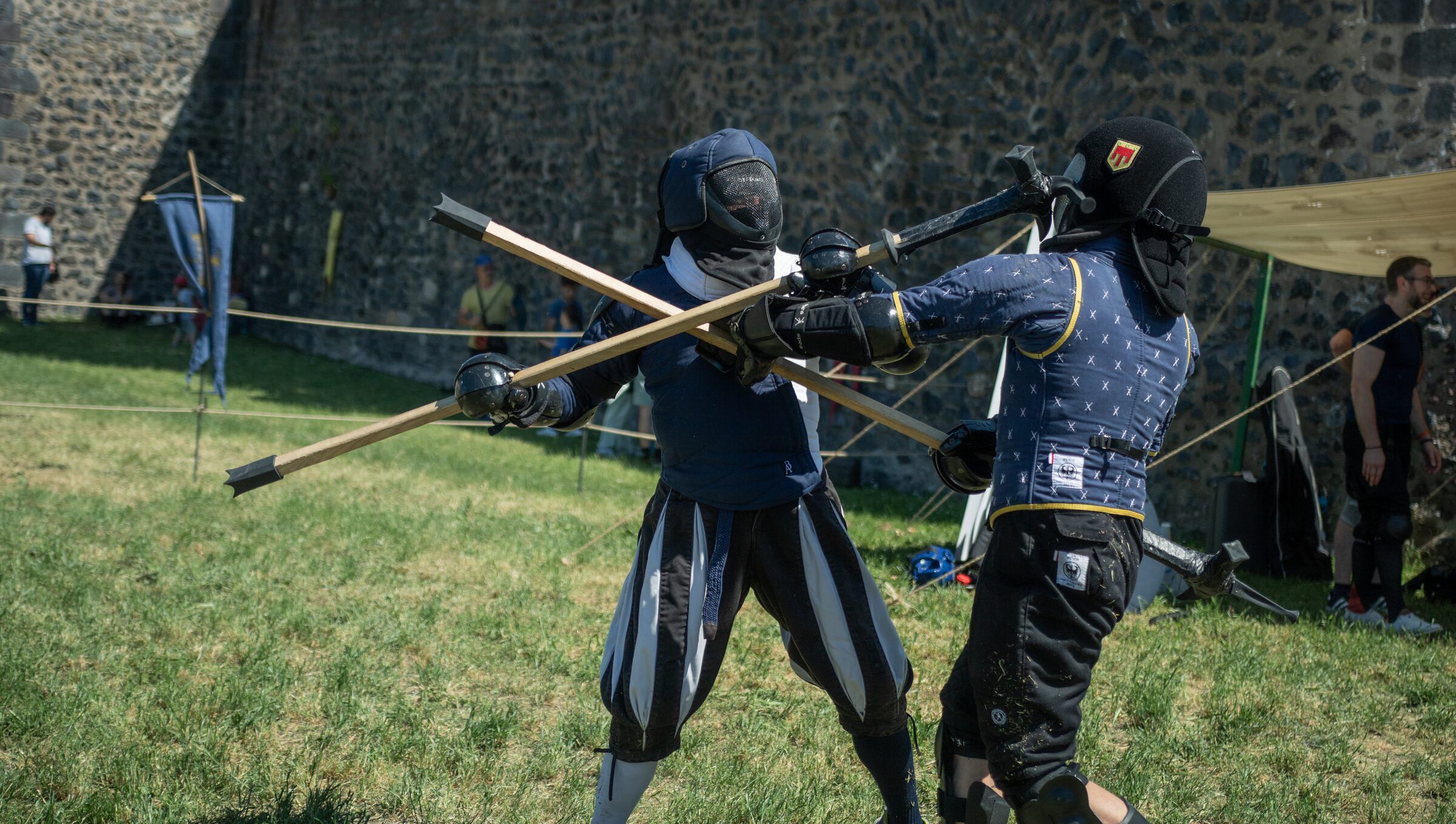 Deux combattants en costume historique croisent leurs hampes devant des remparts de pierre.