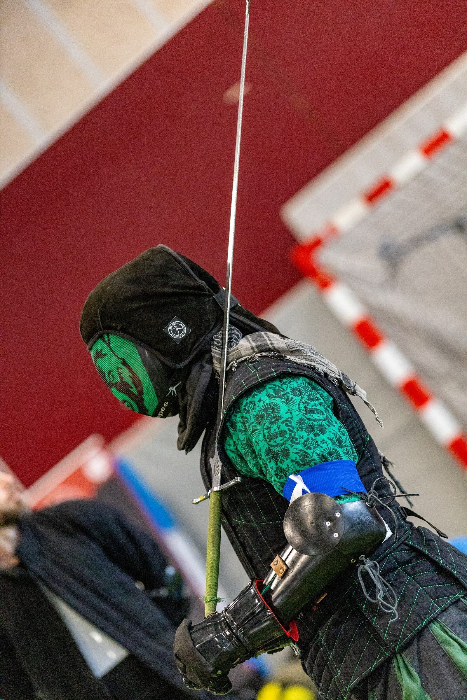 Escrimeur de profil, masque et doublet verts, épée levée devant un mur bordeaux.